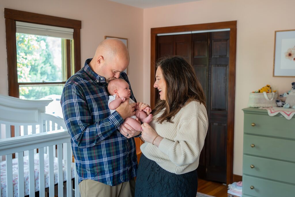 A man and woman are smiling while holding a baby in a nursery room, captured beautifully by Pittsburgh newborn photographers. The room features a crib, dresser, and framed pictures.