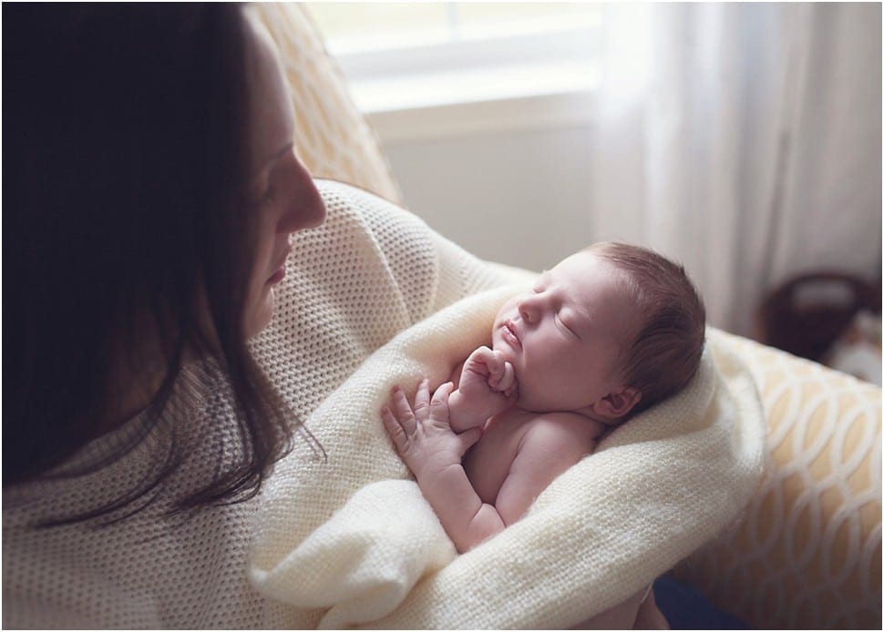 mother in chair in nursery holding newborn baby in window light swaddled in blanket