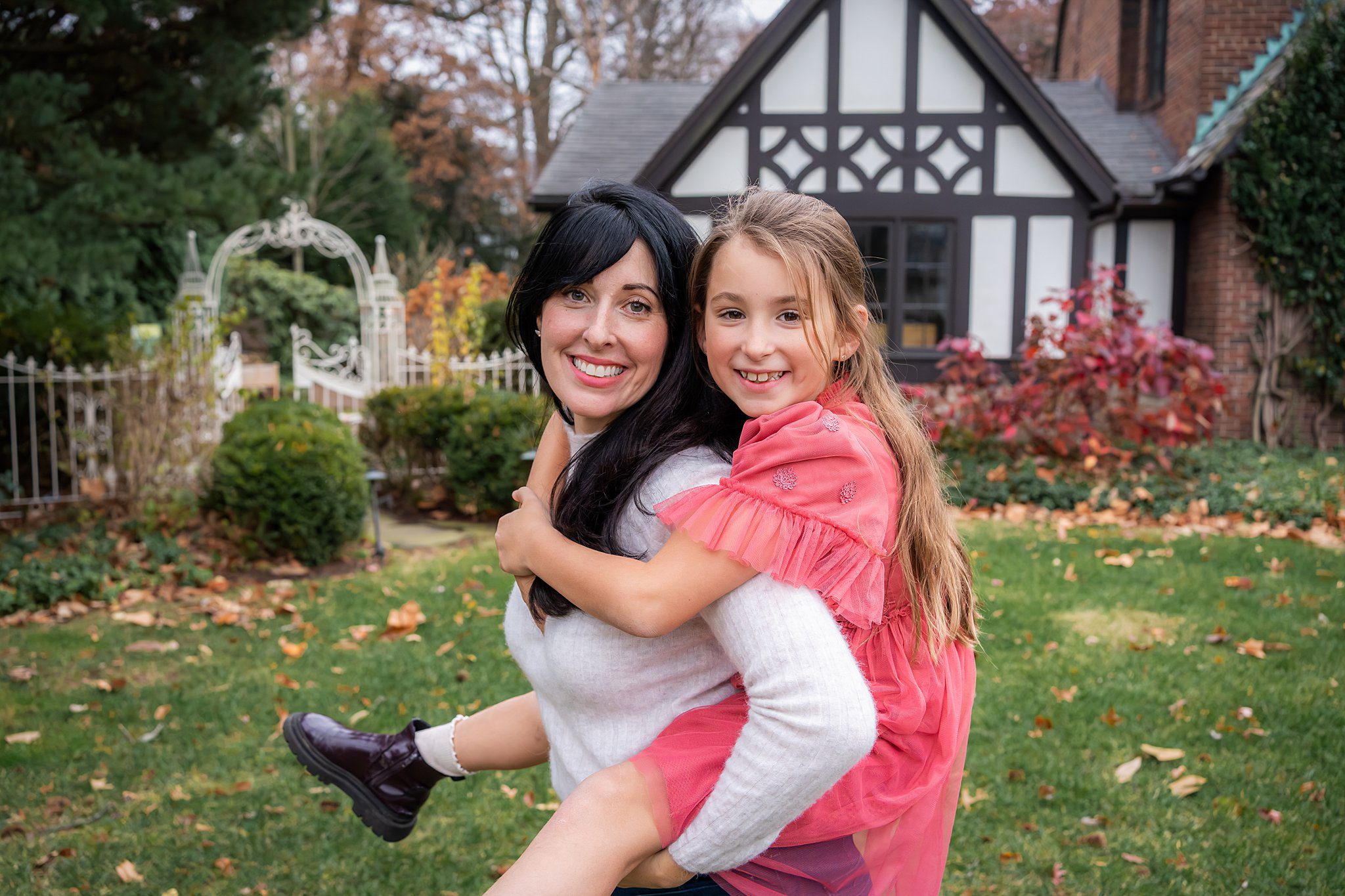 A woman gives a piggyback ride to a smiling young girl in a pink dress, capturing the joy of a family photo session at home in front of their house and garden.