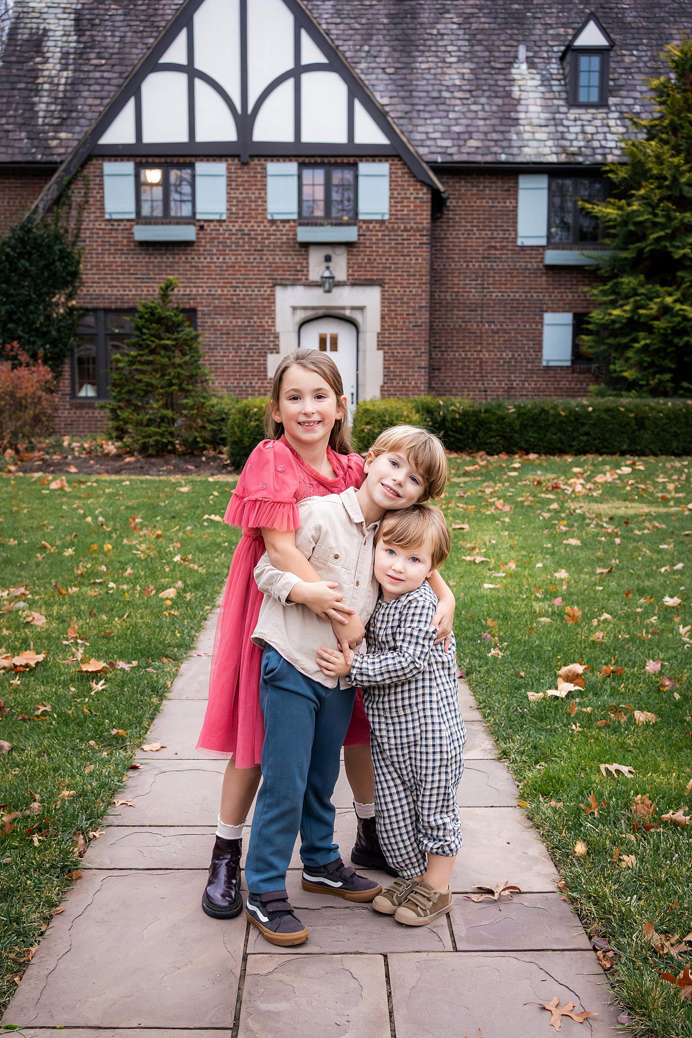 Three children stand on a stone path in front of a large brick house, smiling at the camera during a family photo session at home. The yard is green with scattered autumn leaves.