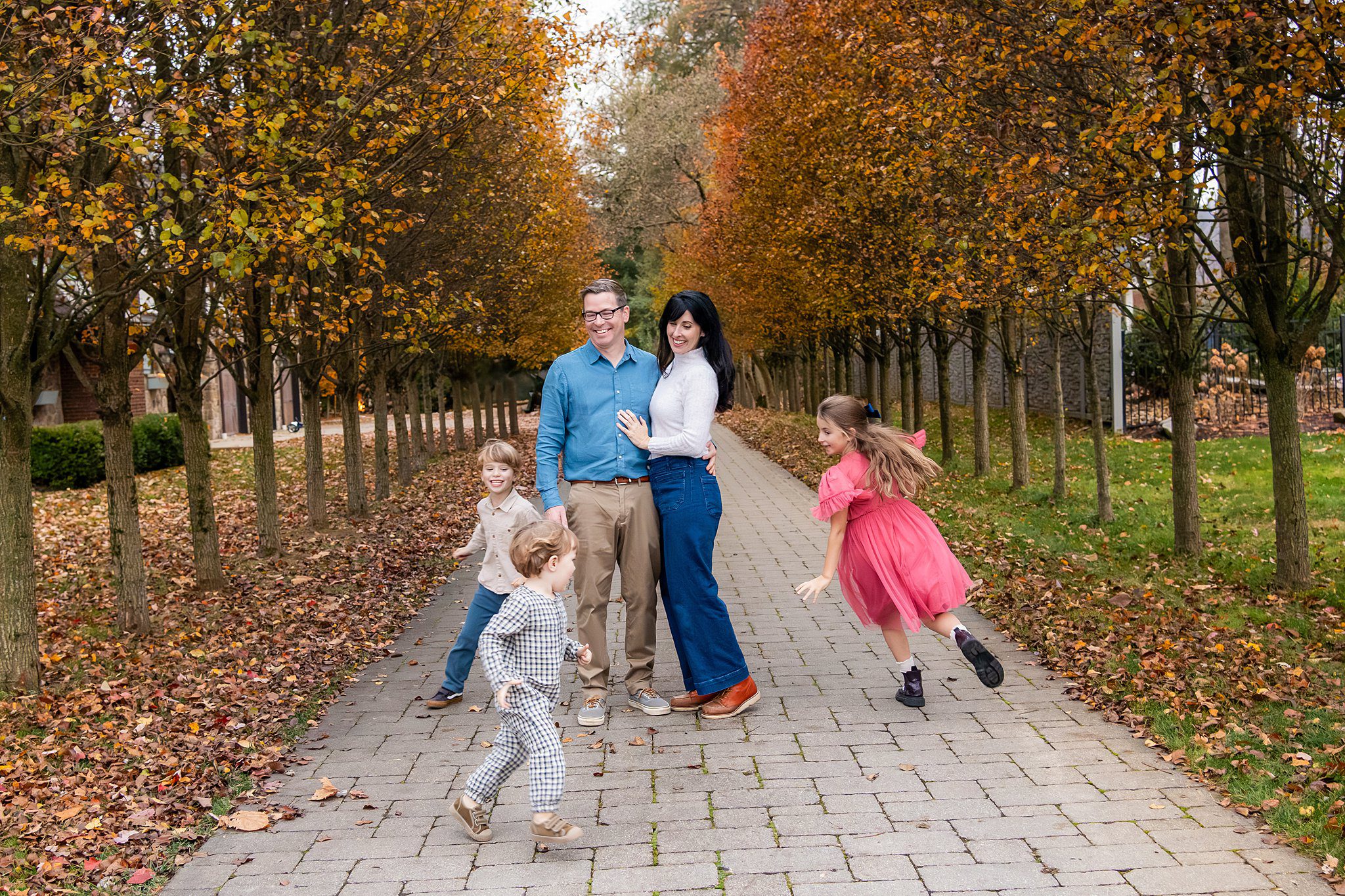 A family photo session at home captures a family of four on a tree-lined path with autumn leaves; two children run and play while two adults pose and smile for the camera.