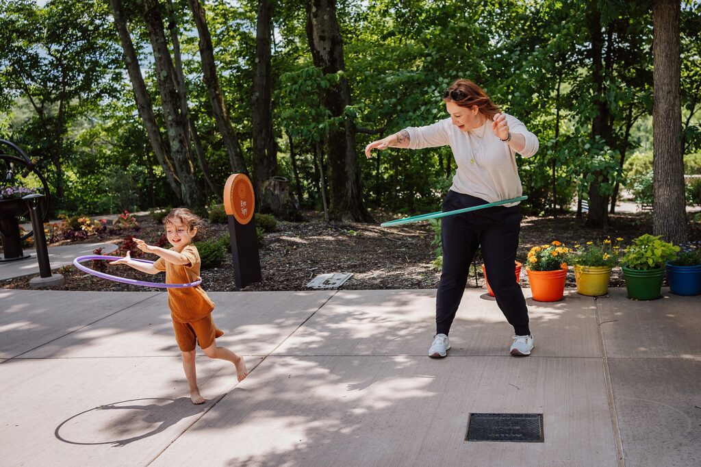 An adult and a child are hula hooping outdoors on a paved area surrounded by trees and colorful flower pots.