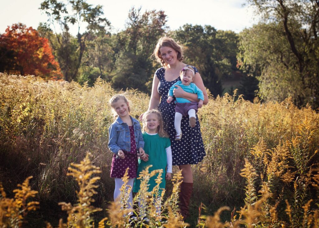 A woman stands in a sunny field with three young children, two girls beside her and a baby in her arms, surrounded by tall yellow wildflowers during a Pittsburgh photo session.
