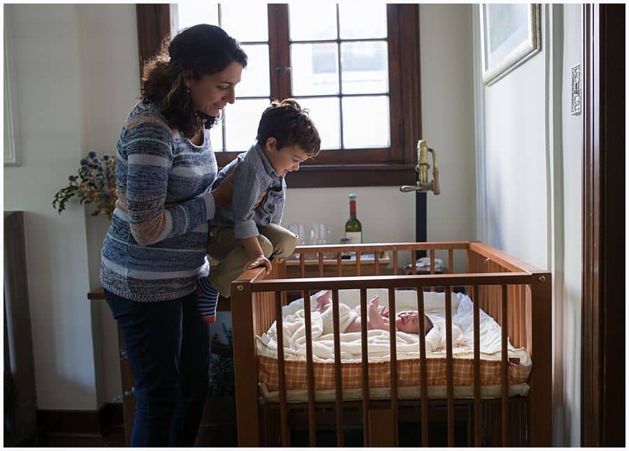 mom and big brother look over baby in square crib