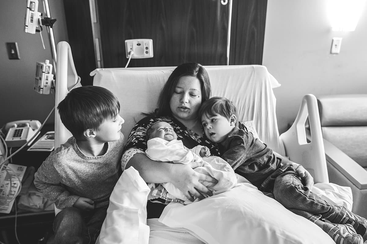 mom holding newborn baby in hospital room bed with older brothers