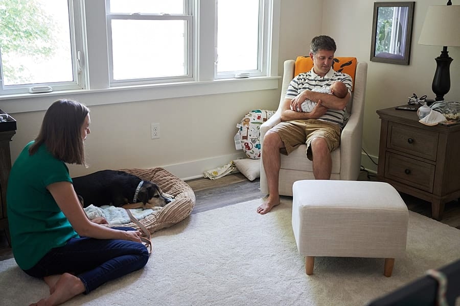 dad looking at newborn son in glider rocker in nursery with mom and dog on floor