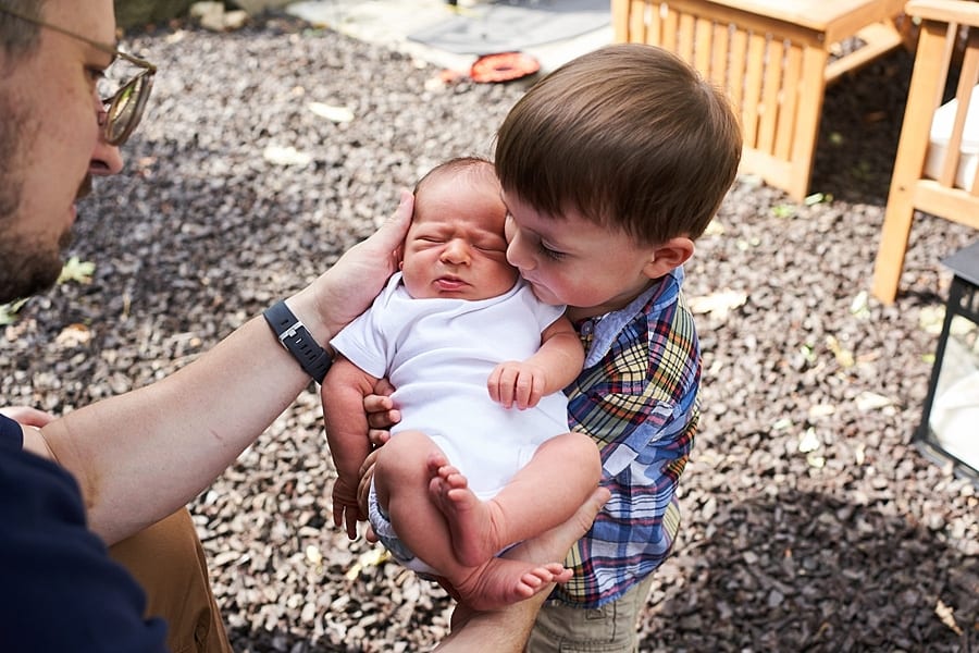 toddler boy holding newborn baby brother  outside with Dad's helping hands for newborn photo session