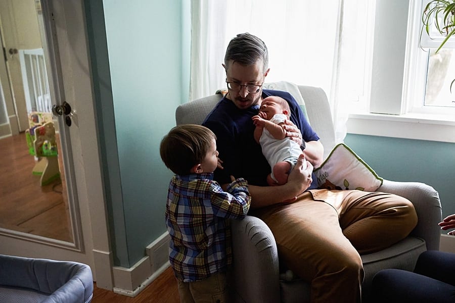 dad holding newborn in rocking chair with big brother looking at baby