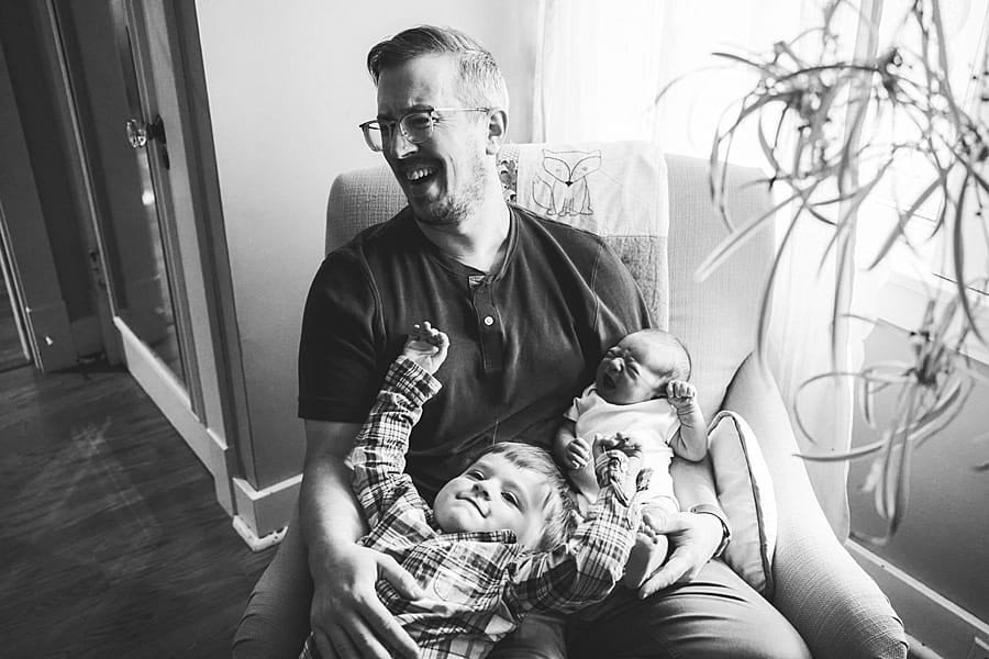 dad laughing while holding newborn and toddler sliding out of his lap