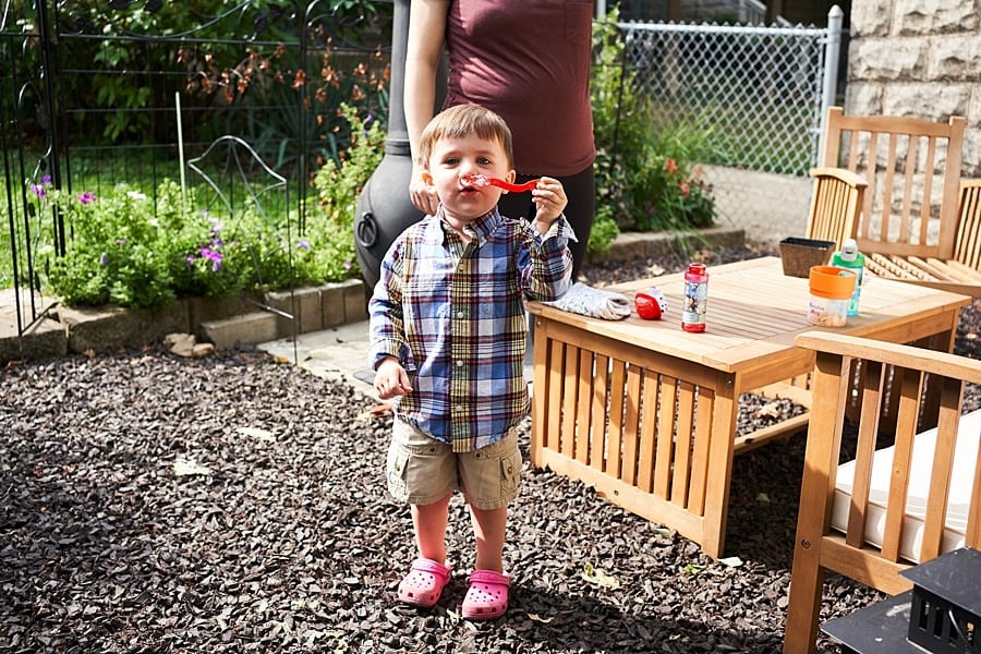 Little boy blowing bubbles outside in yard with his mother standing behind him