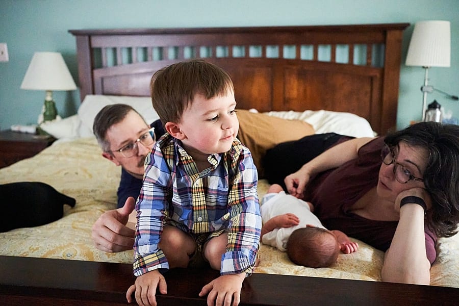 toddler on edge of foot of bed with mom and dad next to him