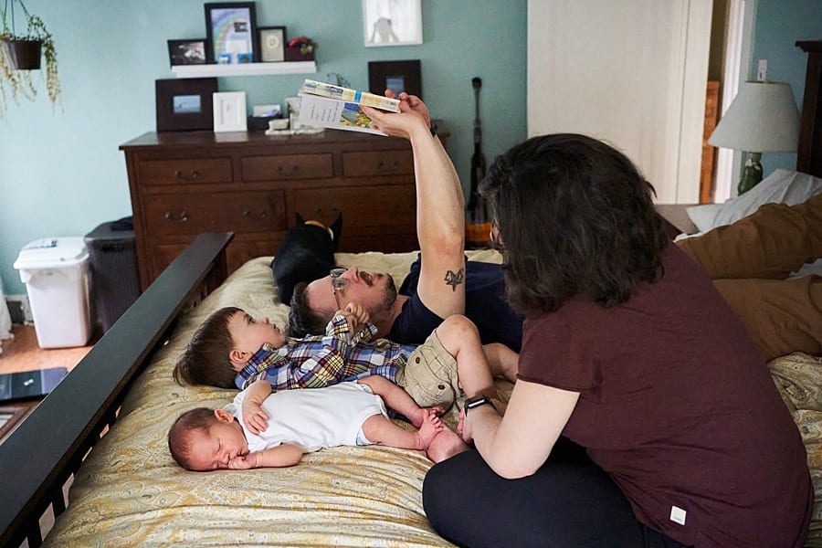 dad and toddler and newborn laying on bed reading a book while mom watches