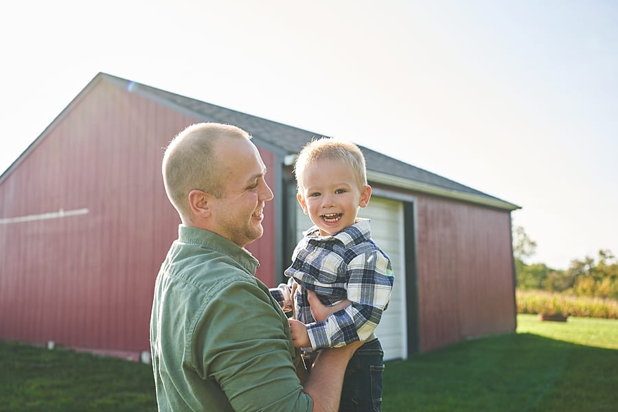 Father holding smiling blonde son in front of a red barn