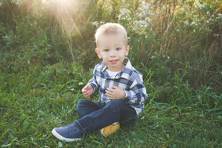 little boy sitting in the grass looking at the camera smiling in the sunlight behind him  with parents for Family Photos with a three year old on the Farm in Beaver County