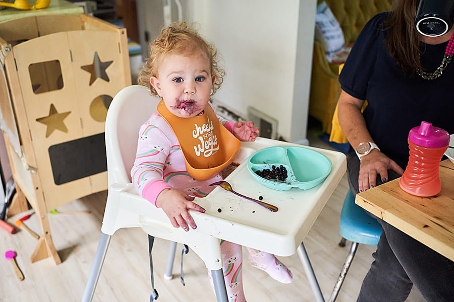 Little girl in her highchair with blueberries all over her face eating breakfast
