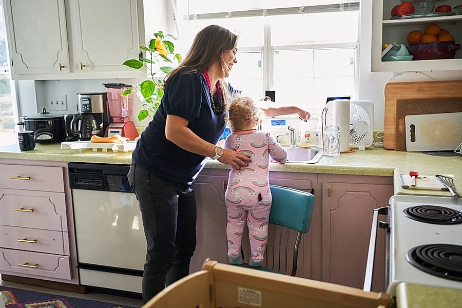Little girl standing on vintage kitchen chair in rainbow pajamas washing her hands at the sink with her mother