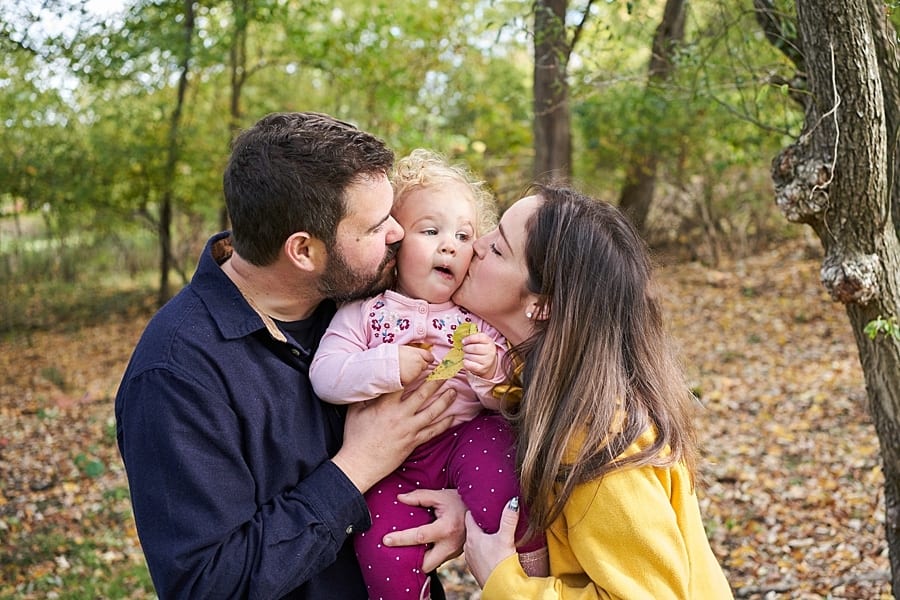 Mother and father kissing toddler girl on cheek in their backyard