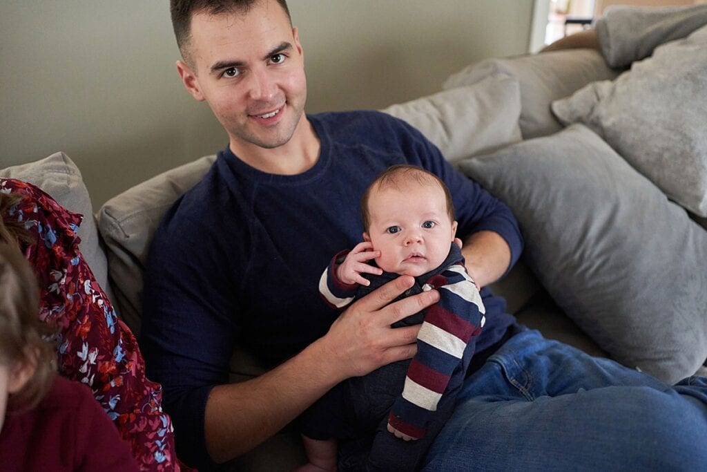 Father sitting on couch holding newborn son while both look into the camera