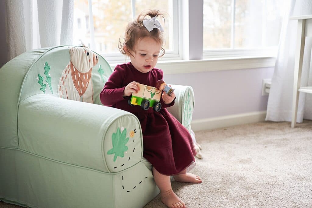 Little girl sitting on a Pottery Barn chair by the window holding a toy