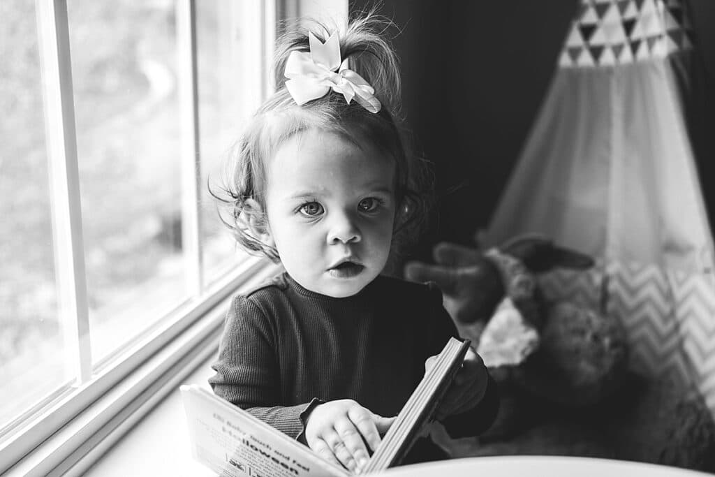 Little girl with a bow on her head looking at a book in front of the window