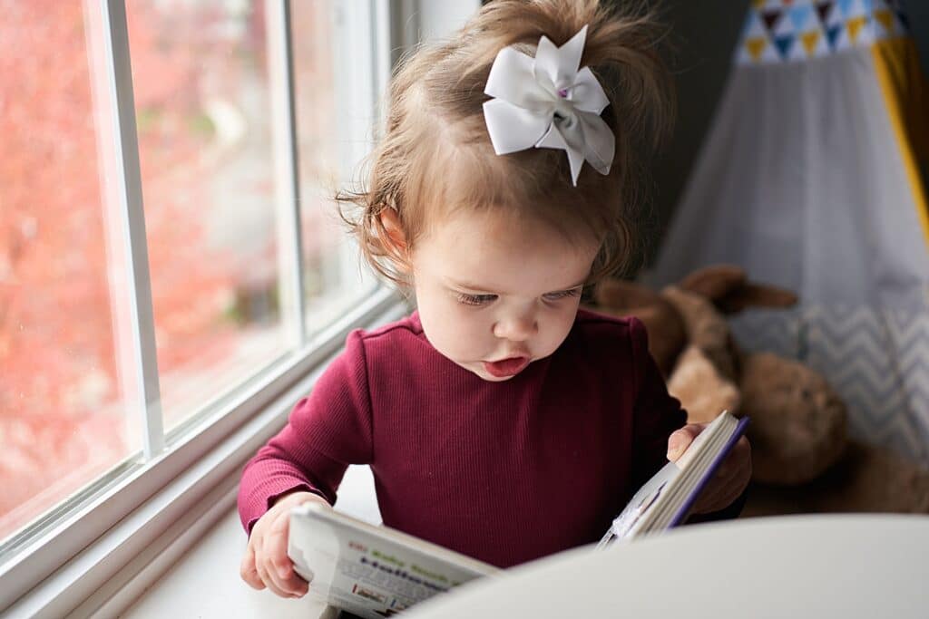 Little girl with a bow on her head reading a book in front of the window
