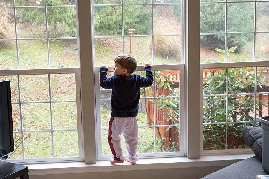 Little boy playing the floor is lava standing in windowsill of large window in living room