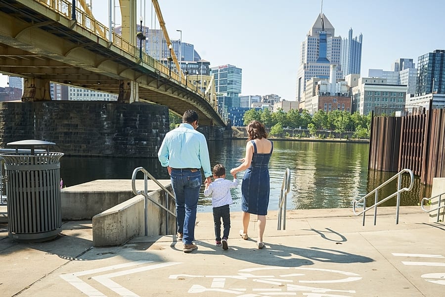 Mother and father holding hands of their son going down the steps in front of PNC park and the Clemente bridge in Pittsburgh with the cityscape in the background