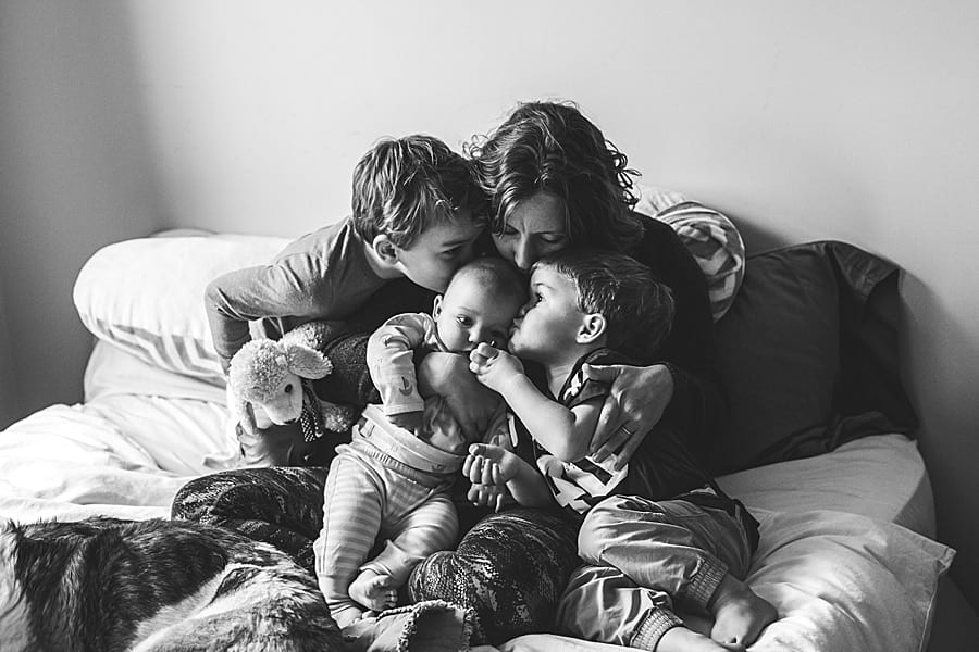 mother on a bed with her three sons and dog kissing the baby