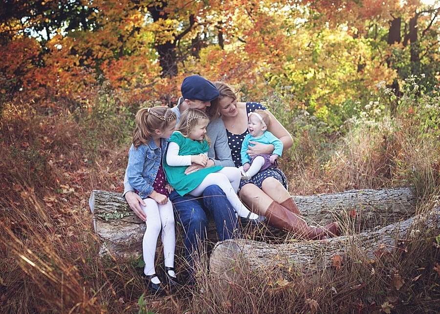 mom dad and three daughters sitting on a log smiling at the baby with fall foliage in the background at fern hollow for a family photo session