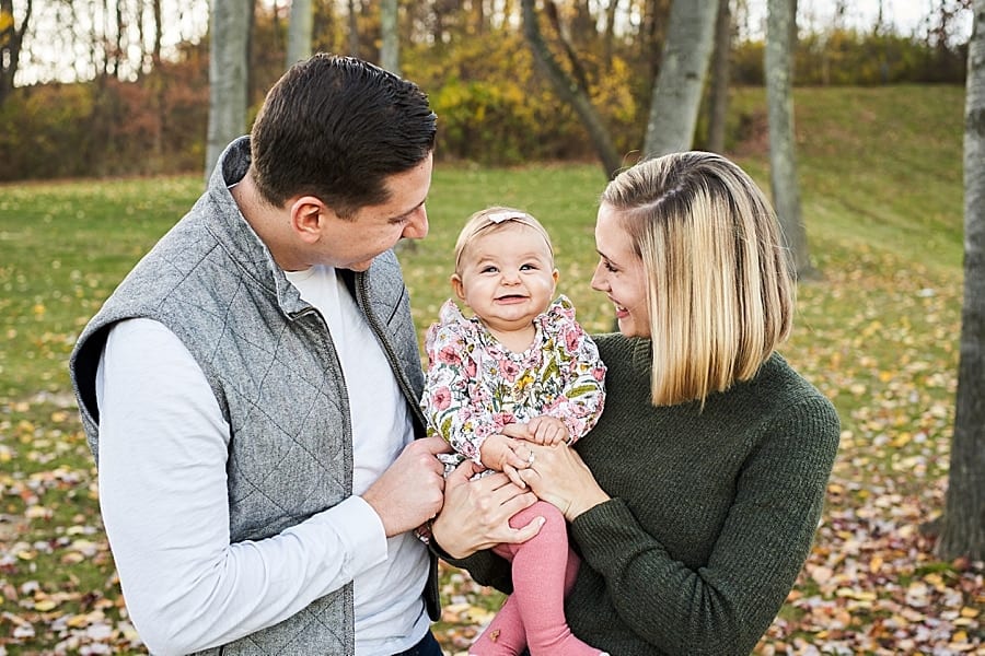 mom dad holding baby girl that is smiling very big with fall foliage in the background for photo session at blueberry hill park cranberry township