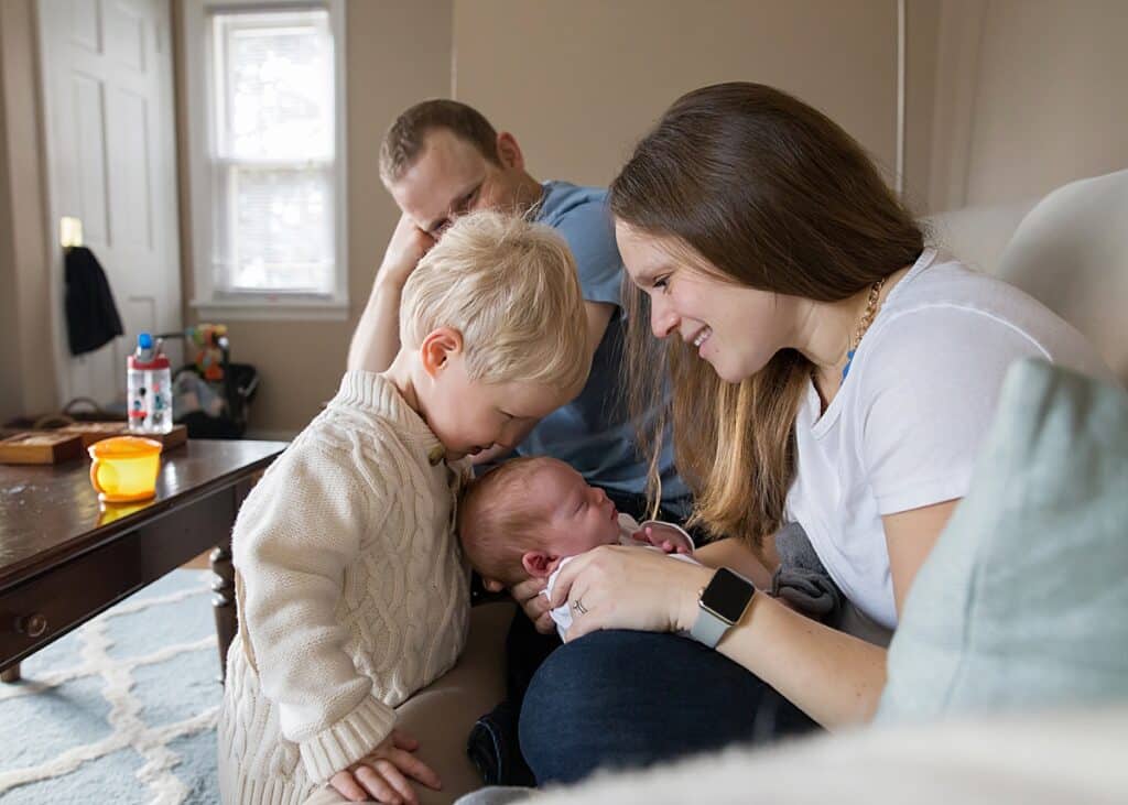 toddler boy kissing newborn baby brother on his head while mom holds baby and dad smile in background of family home How to Find a Newborn Photographer in Pittsburgh
