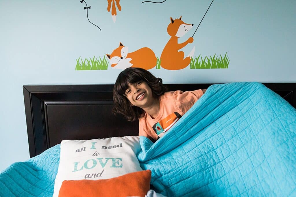 little boy on ned with aqua comforter and fox stickers on the wall smiling at the camera for a family photo session at his home