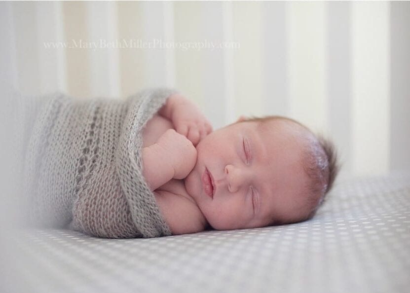 newborn baby wrapped in a gray knit blanket sleeping in crib with hands tucked under chin