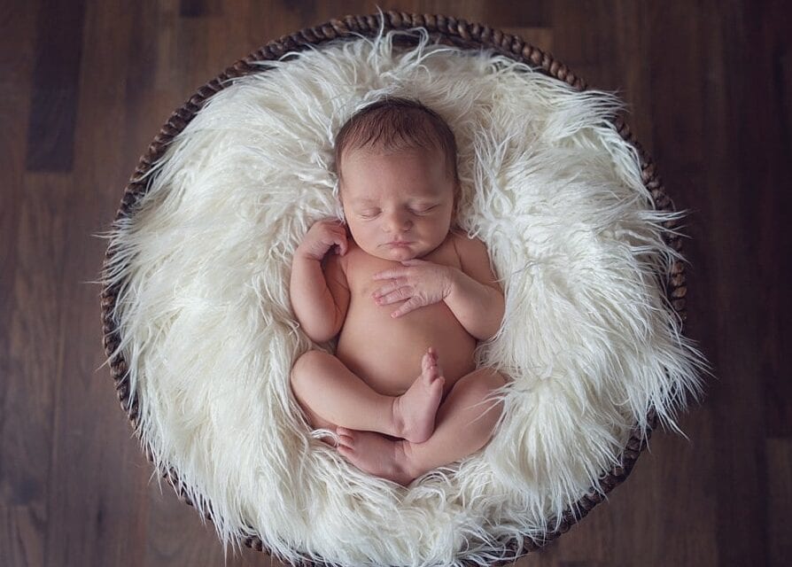 newborn baby laying on back with les curled up hands on chest sleeping on furry white flokati blanket in a basket on a hardwood floor