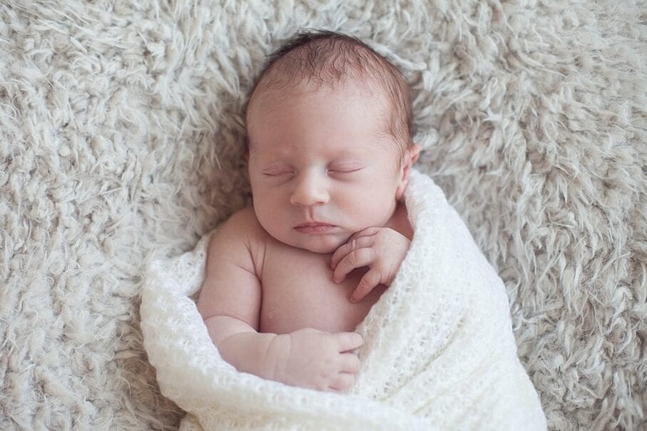 newborn baby sleeping wrapped in a cream knit blanket laying on a gray neutral colored float soft blanket