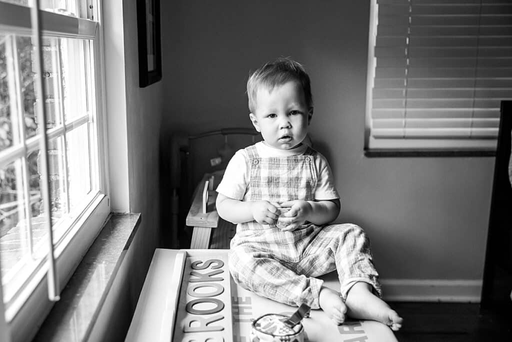 toddler boy sitting on toy chest by a window looking into the camera