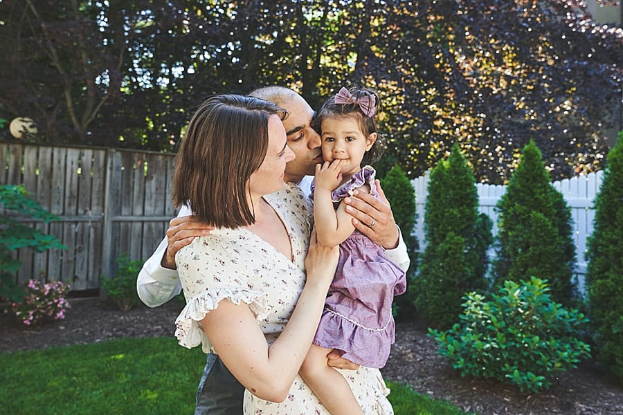 mom and dad kissing daughter in backyard for photo session with Shadyside newborn photographer
