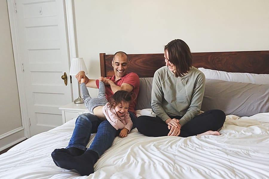 mom dad and little girl playing on the bed in master bedroom of Shadyside home for a family photo session 