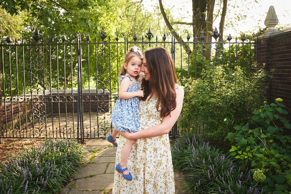 wexford mom holding daughter during a photo session at the walled garden in Mellon Park with black metal gates and brick walls and flowers