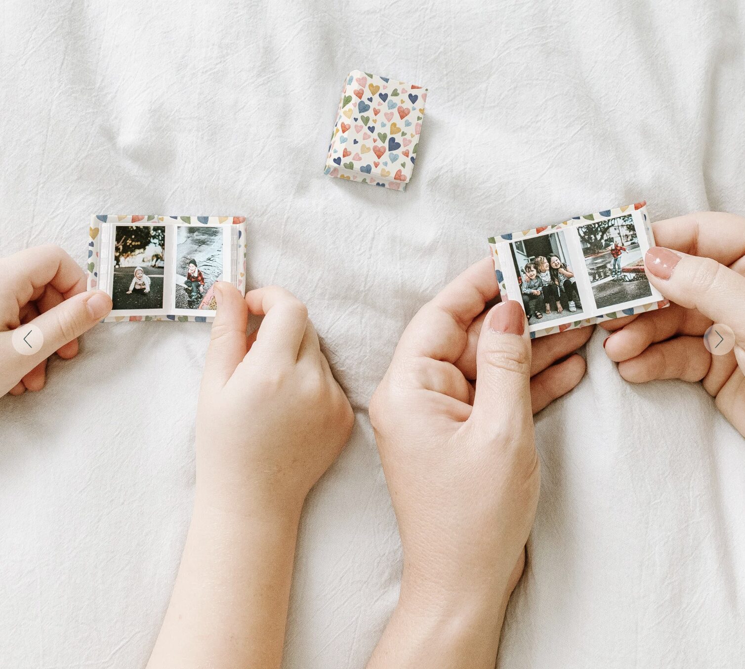 Two people hold small photo strips with three pictures each on a white bed, showing one of the creative ways to get your photos off your phone. A small, heart-patterned box sits above their hands.