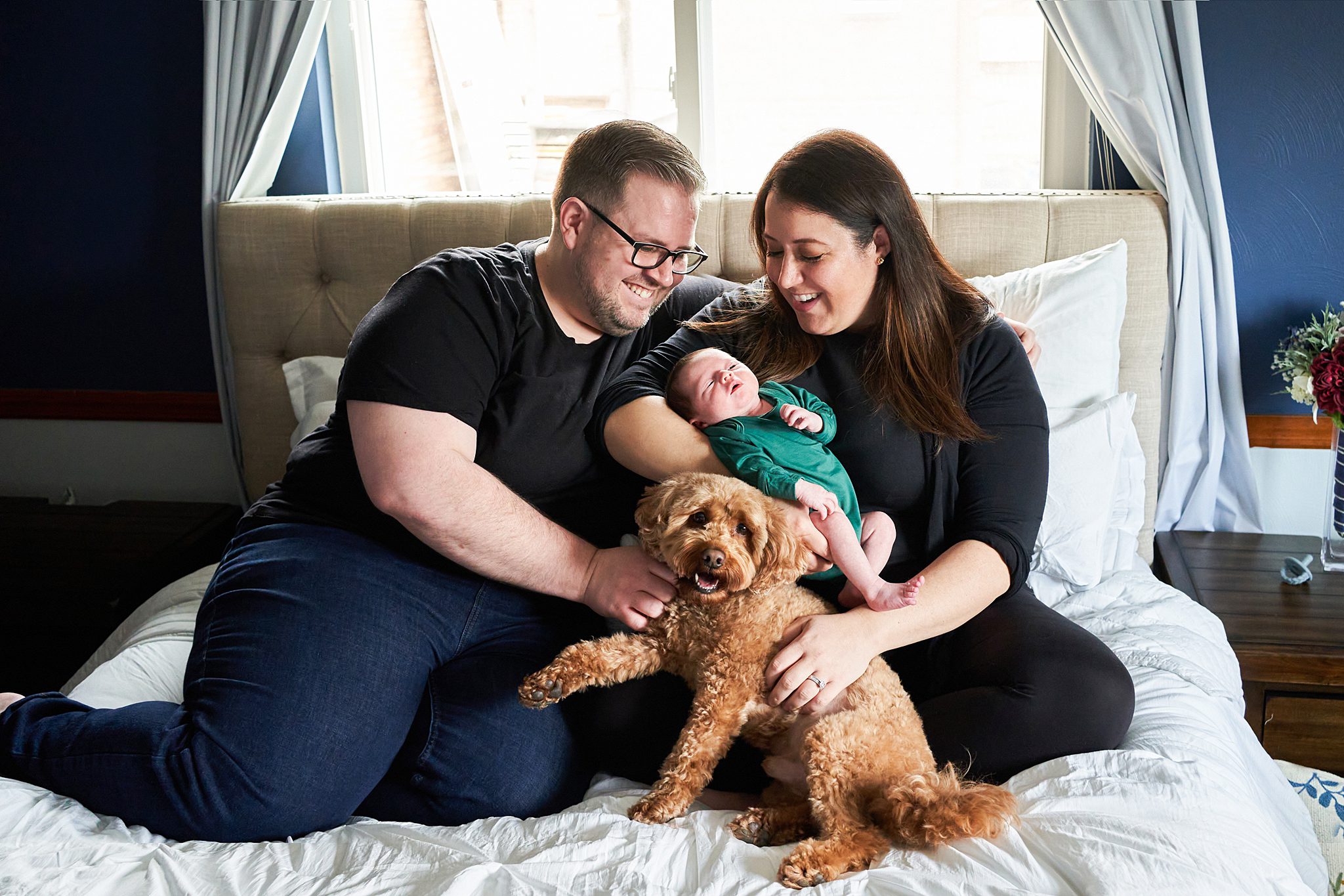 mom and dad on bed with newborn in north hills home with golden doodle dog for a newborn photo session