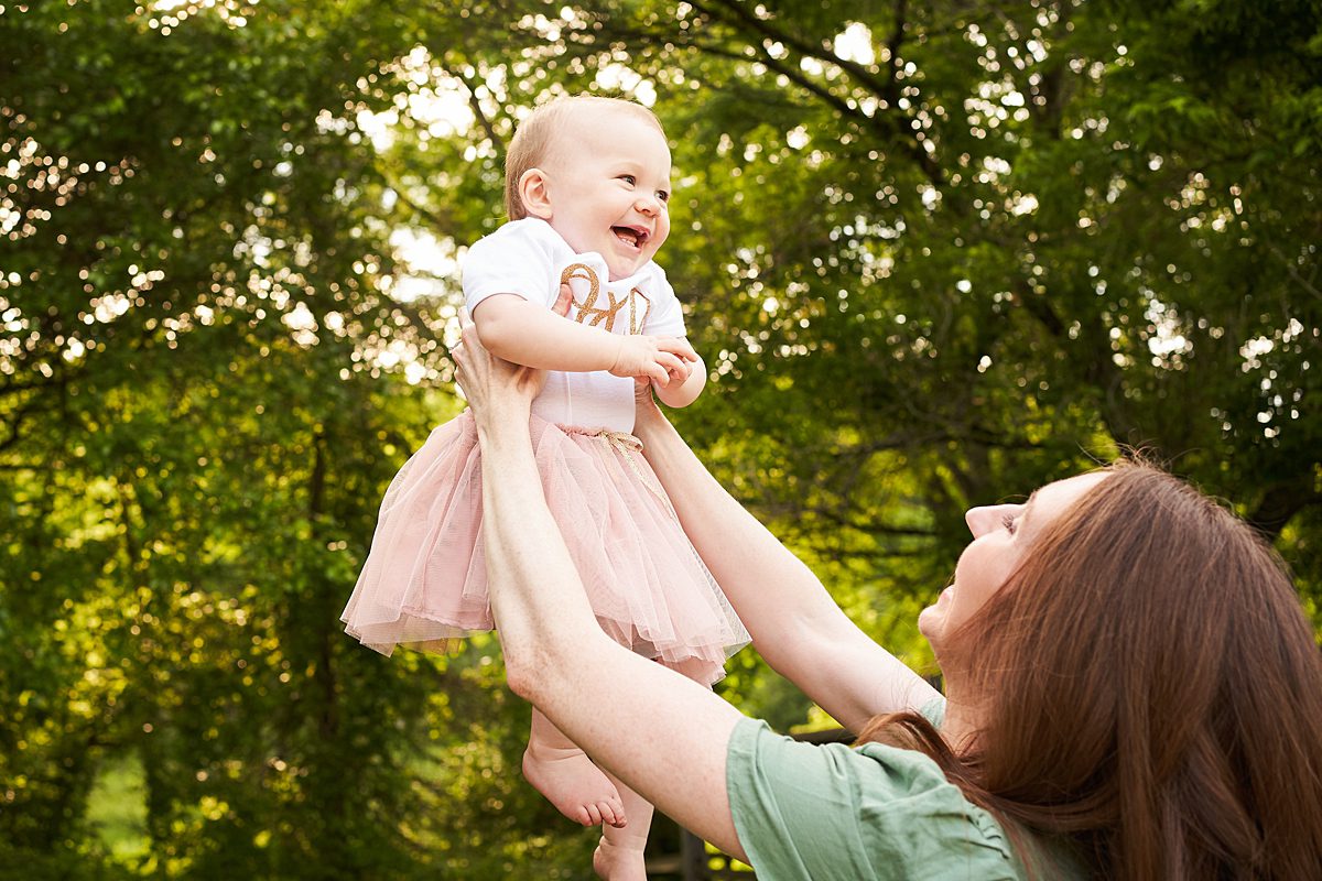 mom holding up baby girl in first birthday outfit in beechwood park in fox chapel for a family photo session 