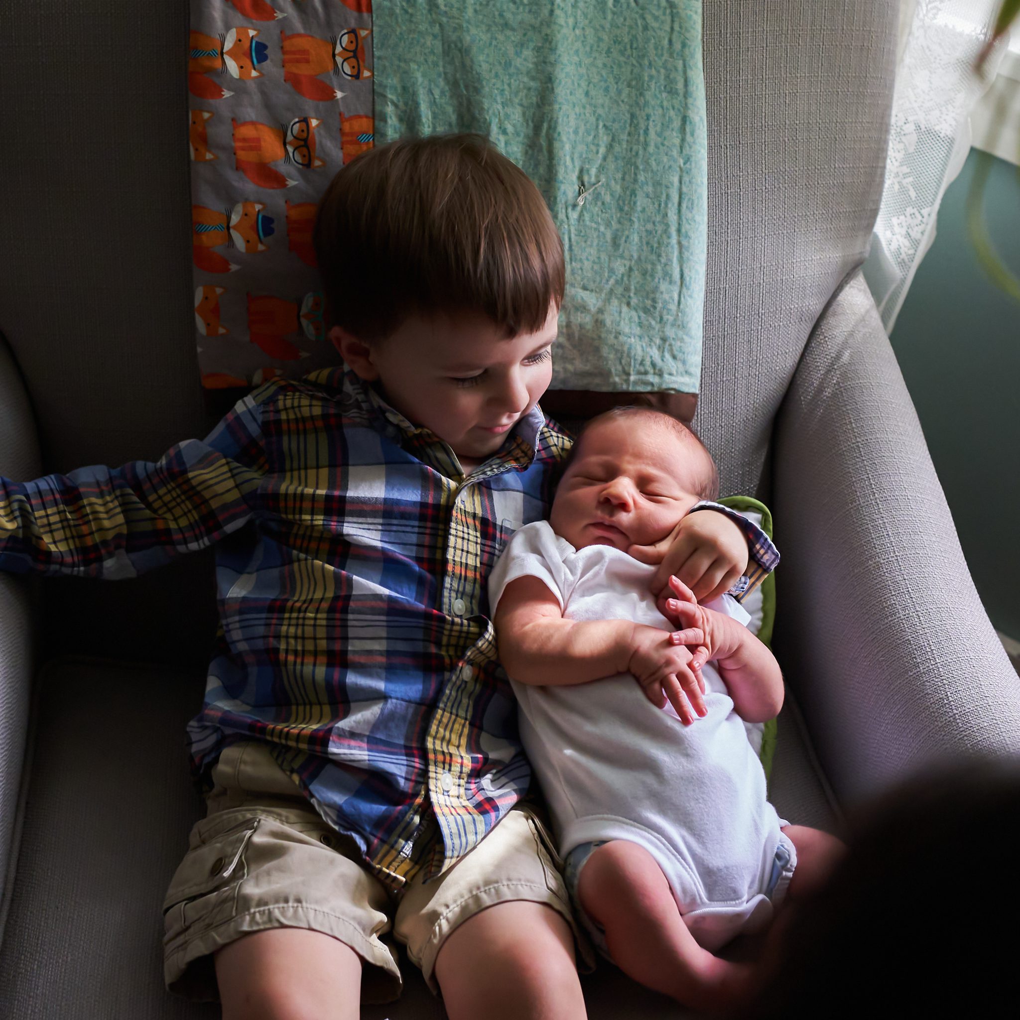 little boy holding newborn baby brother in chair by window inside home