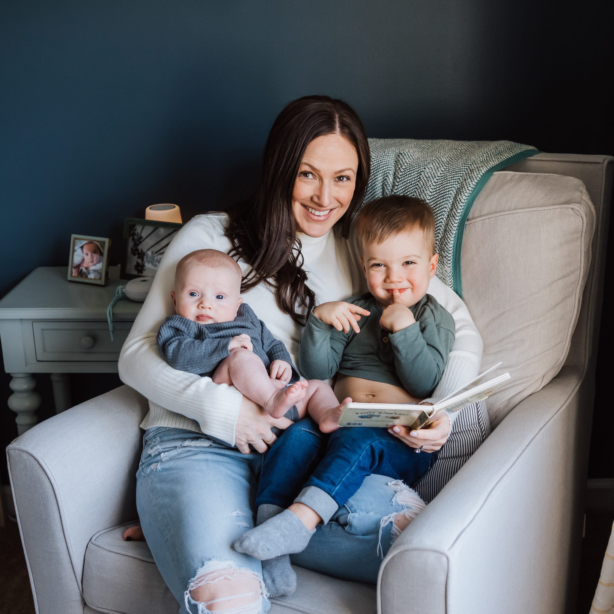 mom reading a book to her toddler son and baby on a chair in his bedroom