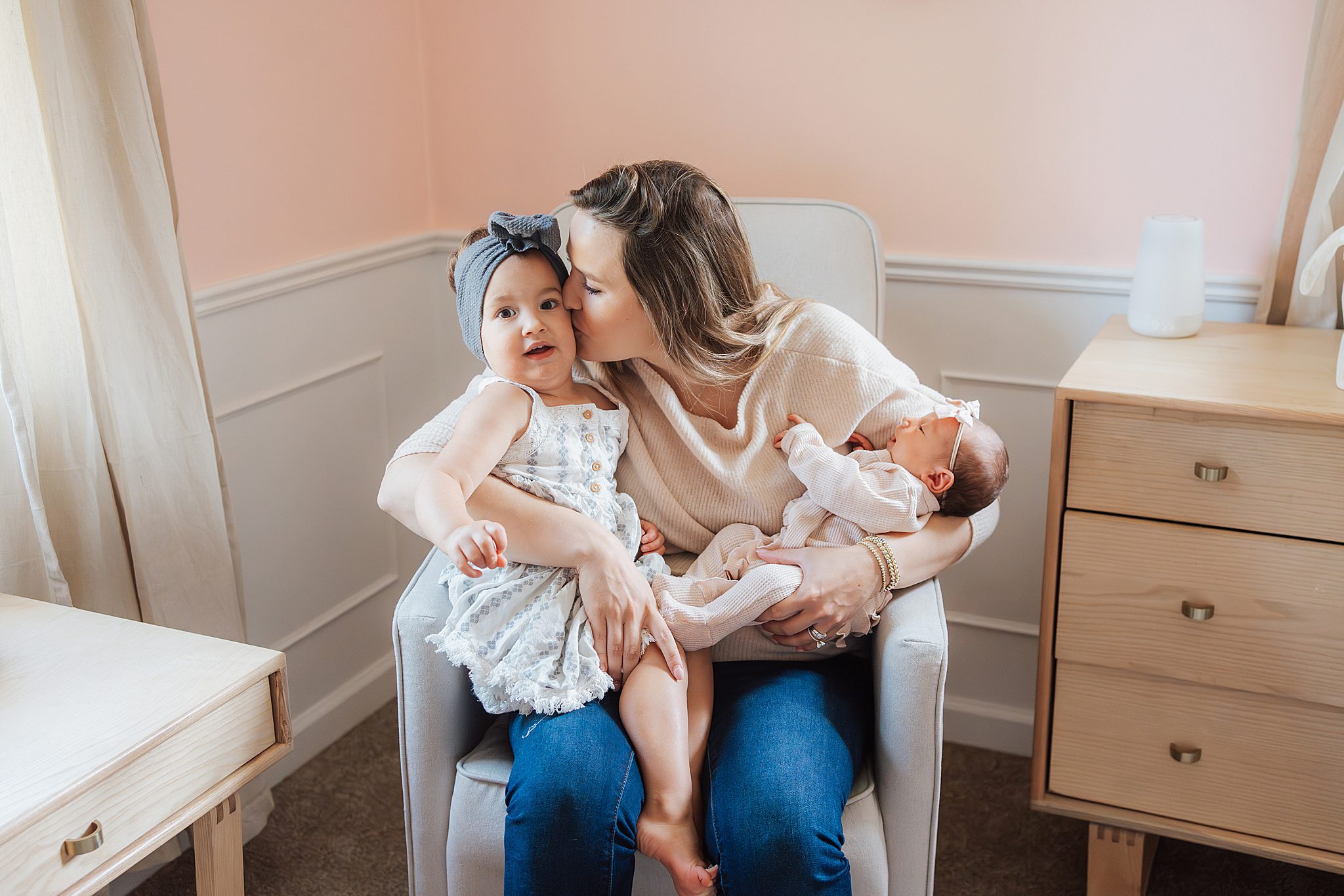 mom kissing daughter while holding newborn on chair in nursery