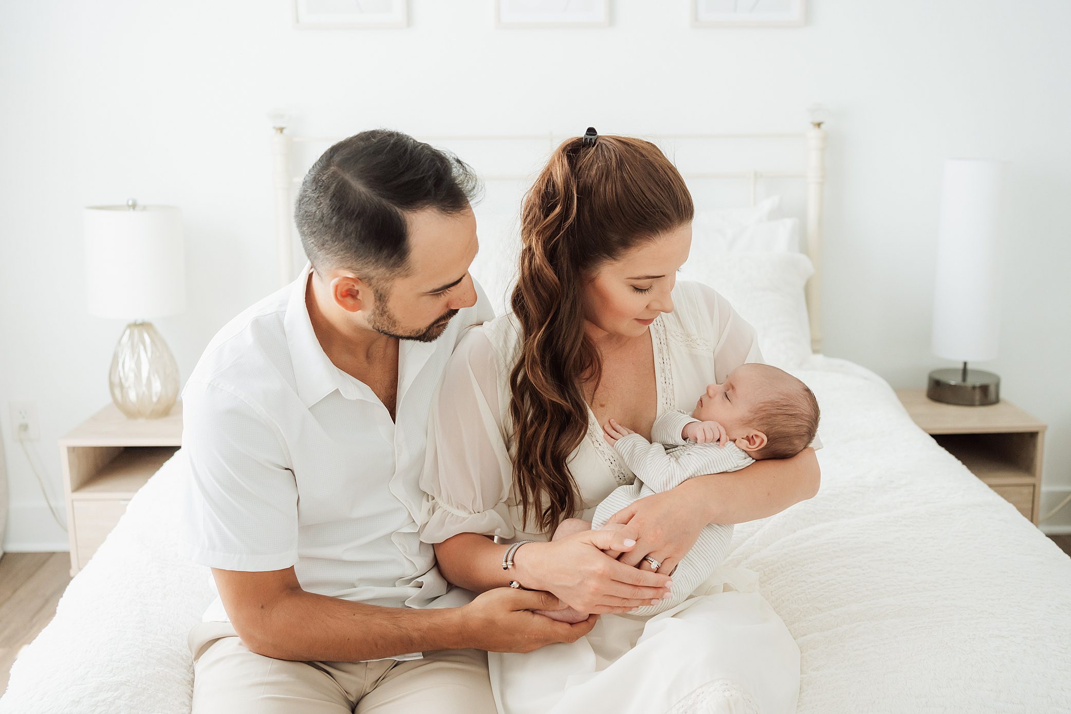 mom and dad in white clothes on a white bed looking at and holding baby
