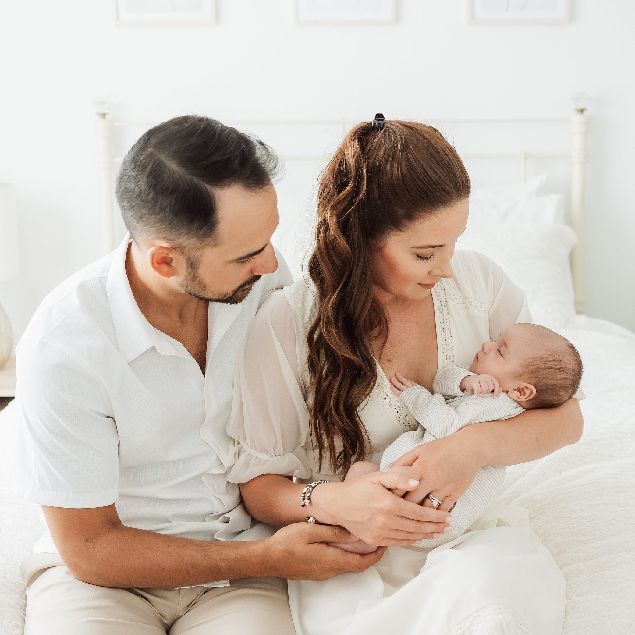 mom, dad in white on a white bed looking at newborn baby