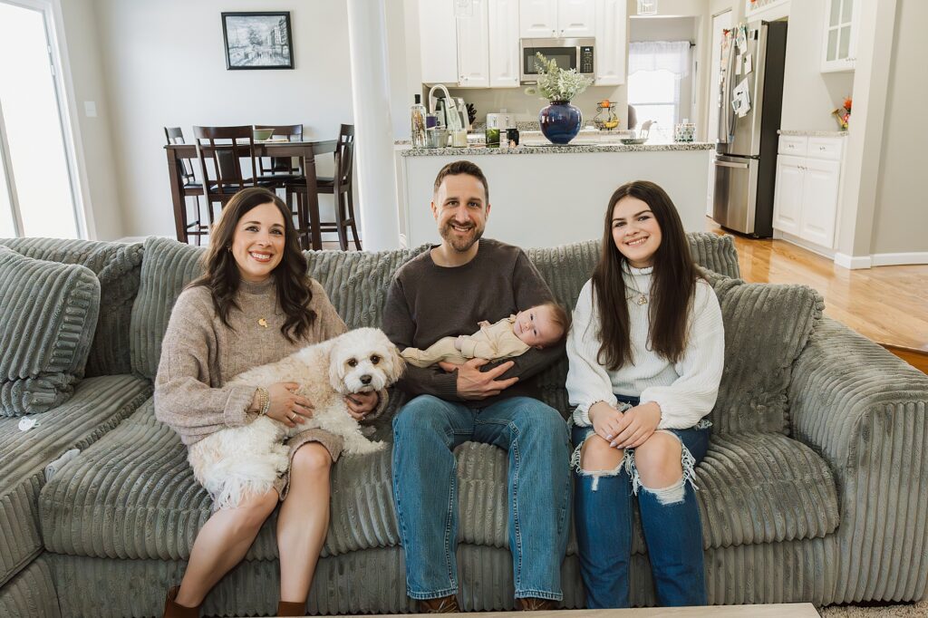 A family with two adults, a teenager, a newborn, and a dog sitting together on a couch in a home with a kitchen in the background, poses for a newborn photographer.