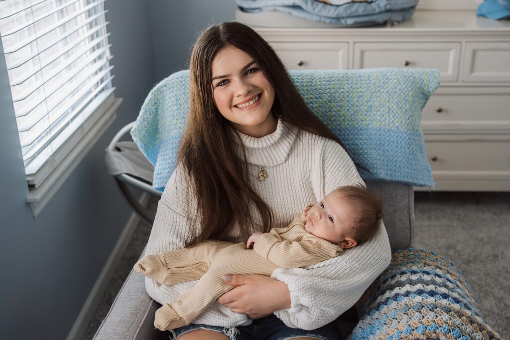 A smiling woman holding a baby while sitting in a cozy room, captured by a newborn photographer.