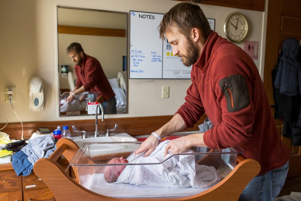 A man is putting a baby in a cradle in hospital room